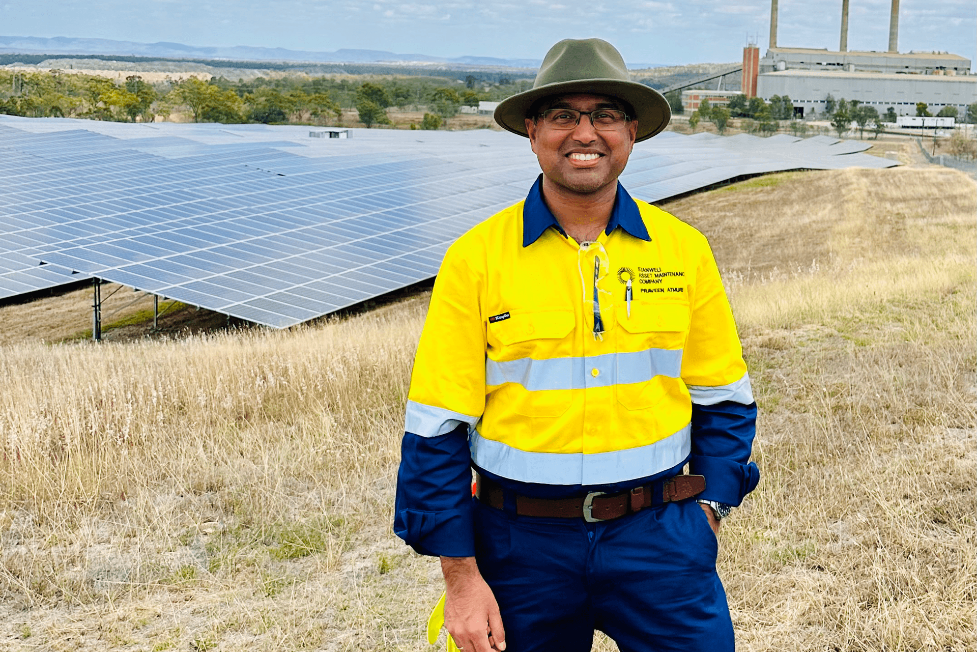 Male in hi vis and a hat smiling at the camera in front of solar panels