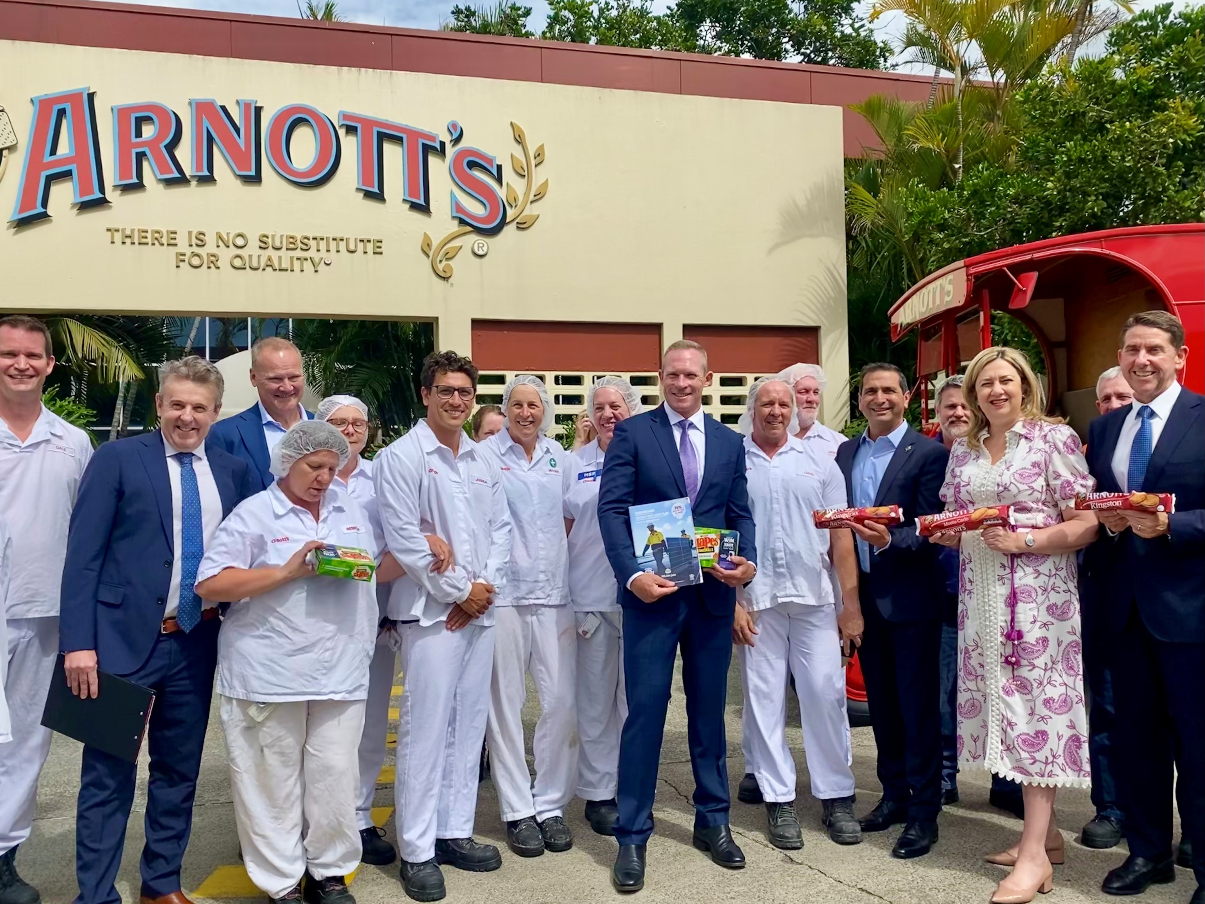 A group of people in corporate wear and factory uniforms pose for a photo with a sign saying Arnotts in the background