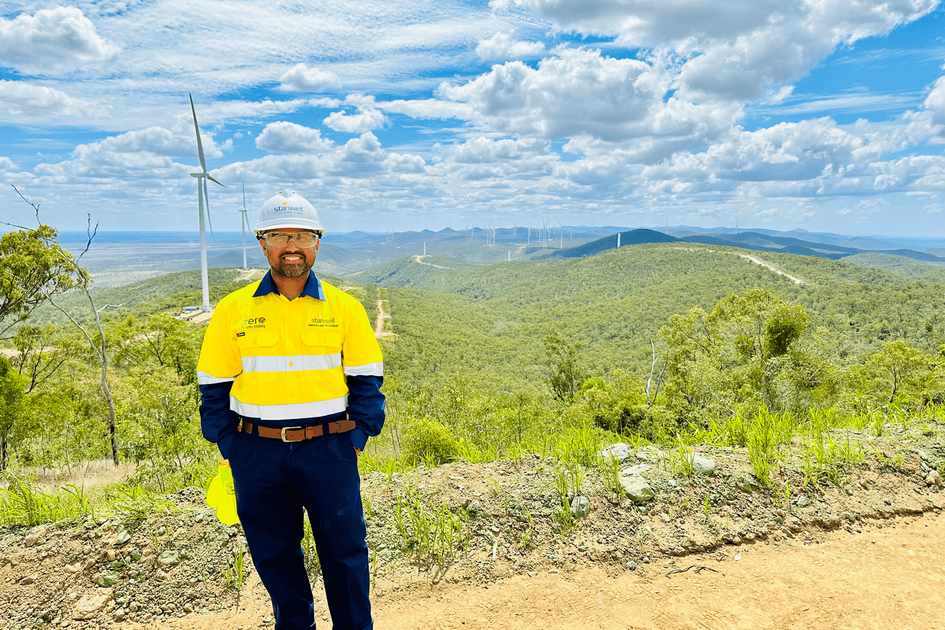 Man smiling at the camera in hi vis and hard hat with a wind farm in the background