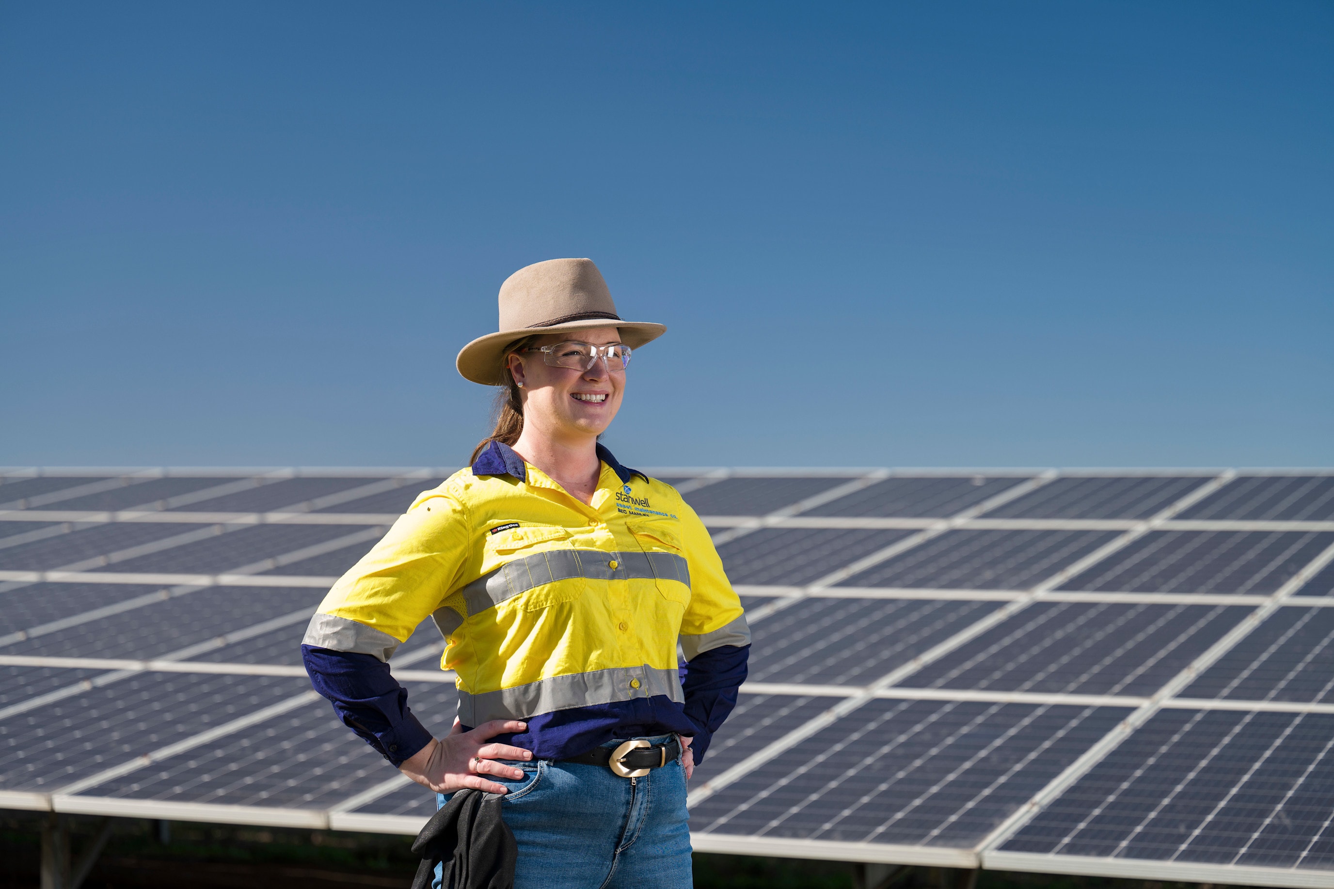 Bec Manley in hi vis, smiling with her hands on her hips in front of solar panels